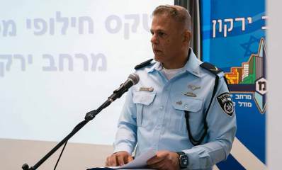 תת ניצב צחי שרעבי מפקד מרחב ירקון עומד לידי דוכן נואמים ולפניו מיקרופון. משמאלו מוקרן הכיתוב: טקס חילופי מפקדים מרחב ירקון, ומימינו הכיתוב: מרחב ירקון וסמל של המרחב.