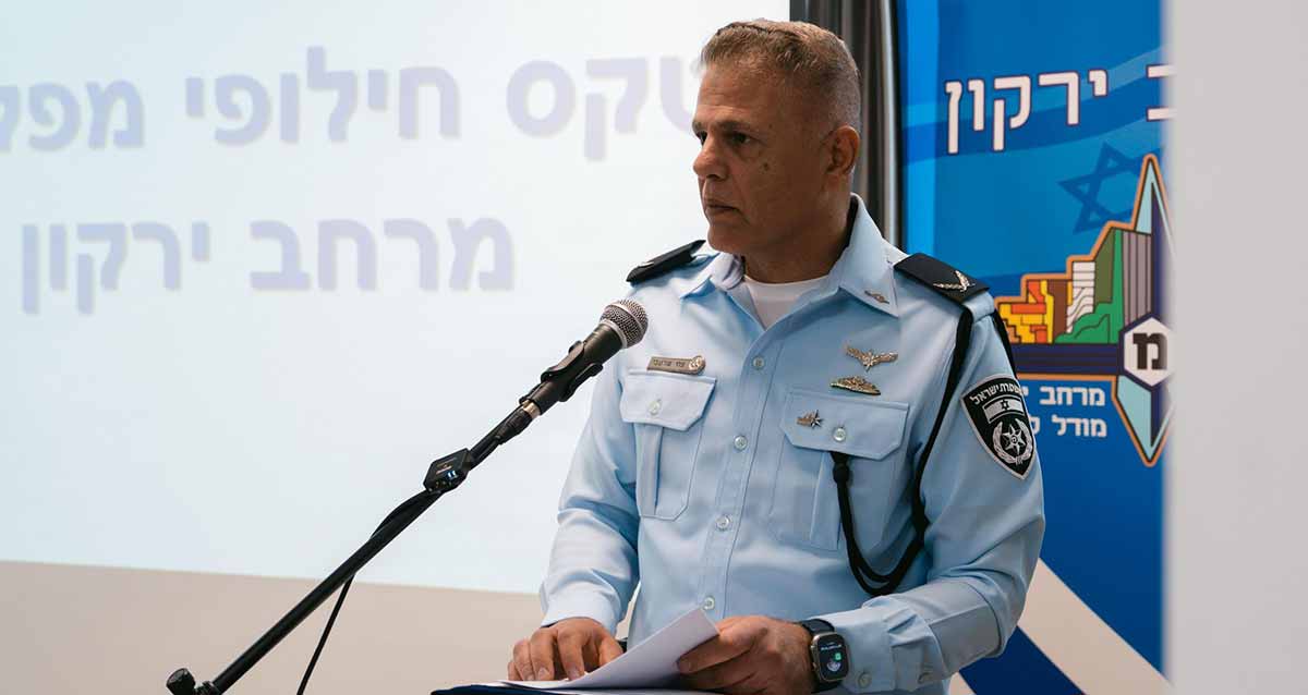 תת ניצב צחי שרעבי מפקד מרחב ירקון עומד לידי דוכן נואמים ולפניו מיקרופון. משמאלו מוקרן הכיתוב: טקס חילופי מפקדים מרחב ירקון, ומימינו הכיתוב: מרחב ירקון וסמל של המרחב.