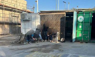 אתר הבנייה בו נתפסו שב"חים. משמאל הבניין עם פיגומים. בחזית מכולות פתוחות. מחוץ למכולה השמאלית יושבים ועומדים שישה שב"חים ופניהם מטושטשות.