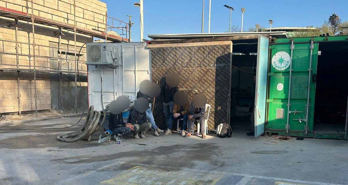 אתר הבנייה בו נתפסו שב"חים. משמאל הבניין עם פיגומים. בחזית מכולות פתוחות. מחוץ למכולה השמאלית יושבים ועומדים שישה שב"חים ופניהם מטושטשות.
