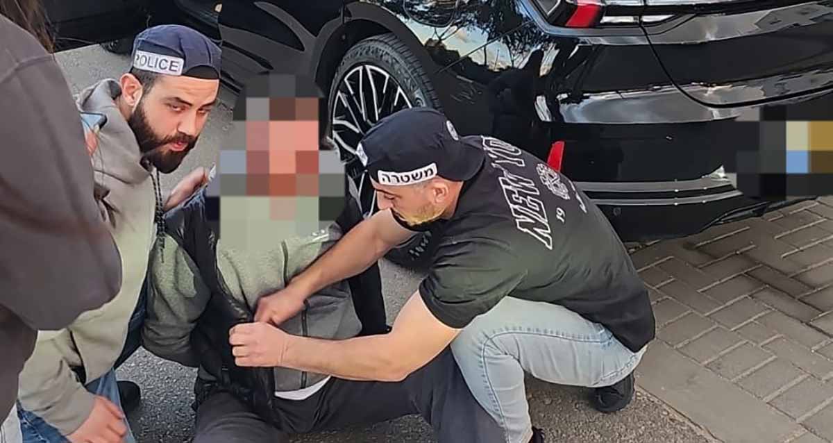 החשוד בדקירה בתחנת הדלק בגלילות. שני שוטרים עושים חיפוש על החשוד היושב על המדרכה.