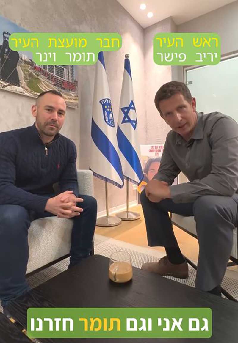 צילום מתוך האינסטגרם של יריב פישר. יריב יושב על כורסא לידו חבר מועצת העיר תומר וינר. הכיתוב בתחתית התמונה גם אני וגם תומר חזרנו. על השולחן לפניהם מונחת כוס עם קפה. מאחוריהם דגלי ישראל ועיריית הרצליה. מעט מוסתר מאחוריהם השלט לשחרור עומר שם טוב.