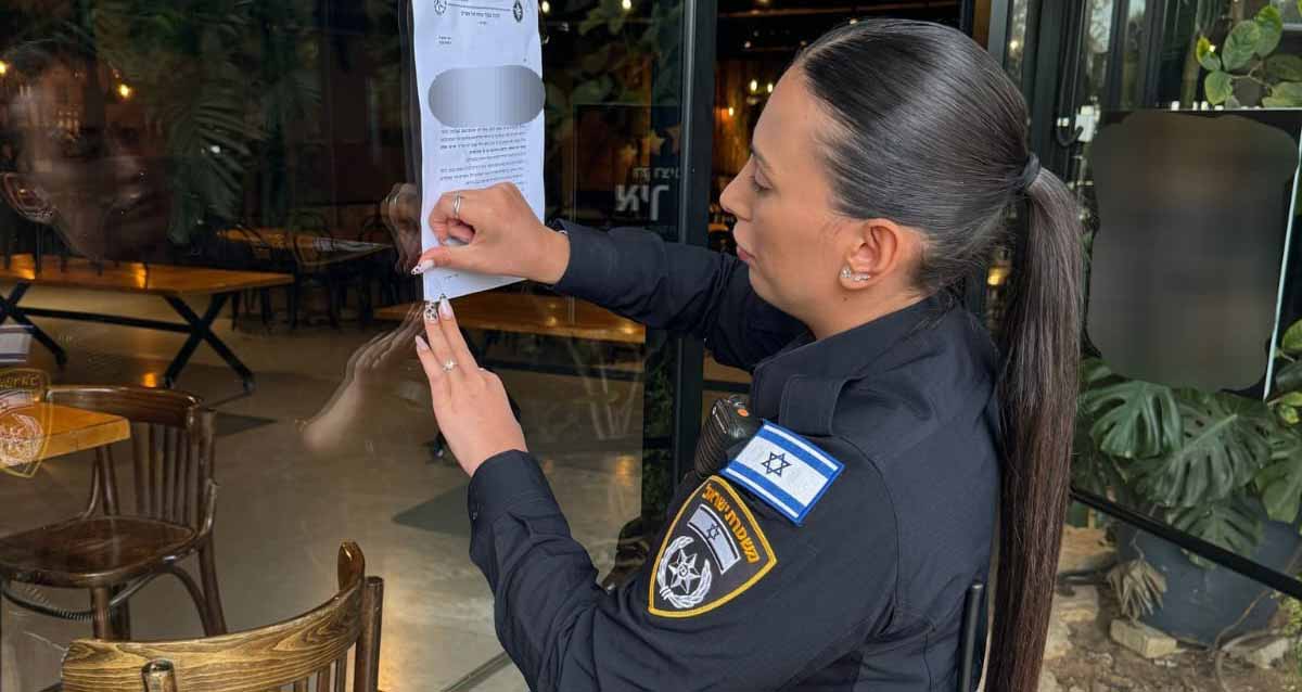 צו סגירה לאולם אירועים ברמת השרון. שוטרת מדביקה את הצו על קיר הזכוכית של האולם. מעבר לזכוכית נראה פנים האולם עם שולחנות וכסאות.