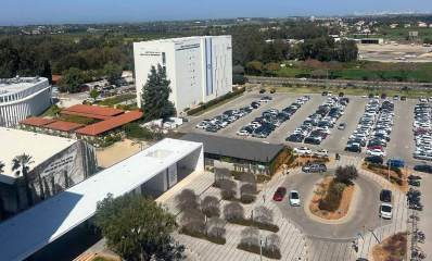 הכניסה אל קמפוס אוניברסיטת רייכמן. מבט על. מימין מגרש חנייה גדול. משמאלו בניין לבן גדול.. בשמאל התמונה מבנה חד קומתי עם גג חום, ולידו בניין מעוגל. בחזית התמונה מרחב הכניסה לקמפוס עם כיכר ועצים, ובשמאל מבנה הכניסה החד קומתי בצבע לבן.