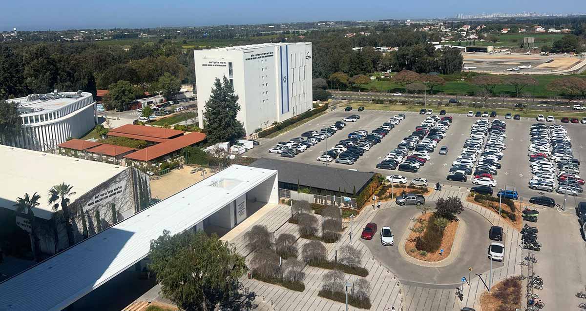 הכניסה אל קמפוס אוניברסיטת רייכמן. מבט על. מימין מגרש חנייה גדול. משמאלו בניין לבן גדול.. בשמאל התמונה מבנה חד קומתי עם גג חום, ולידו בניין מעוגל. בחזית התמונה מרחב הכניסה לקמפוס עם כיכר ועצים, ובשמאל מבנה הכניסה החד קומתי בצבע לבן.