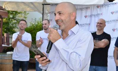 צוות גיאומטריקס רעננה. אדם עם מיקרופון וטלפון ביד עומד לפני קבוצה של אנשים. מאחוריהם על הקיר יריעת בד עם סמל החברה ושמה באנגלית: GEOMATRIX.
