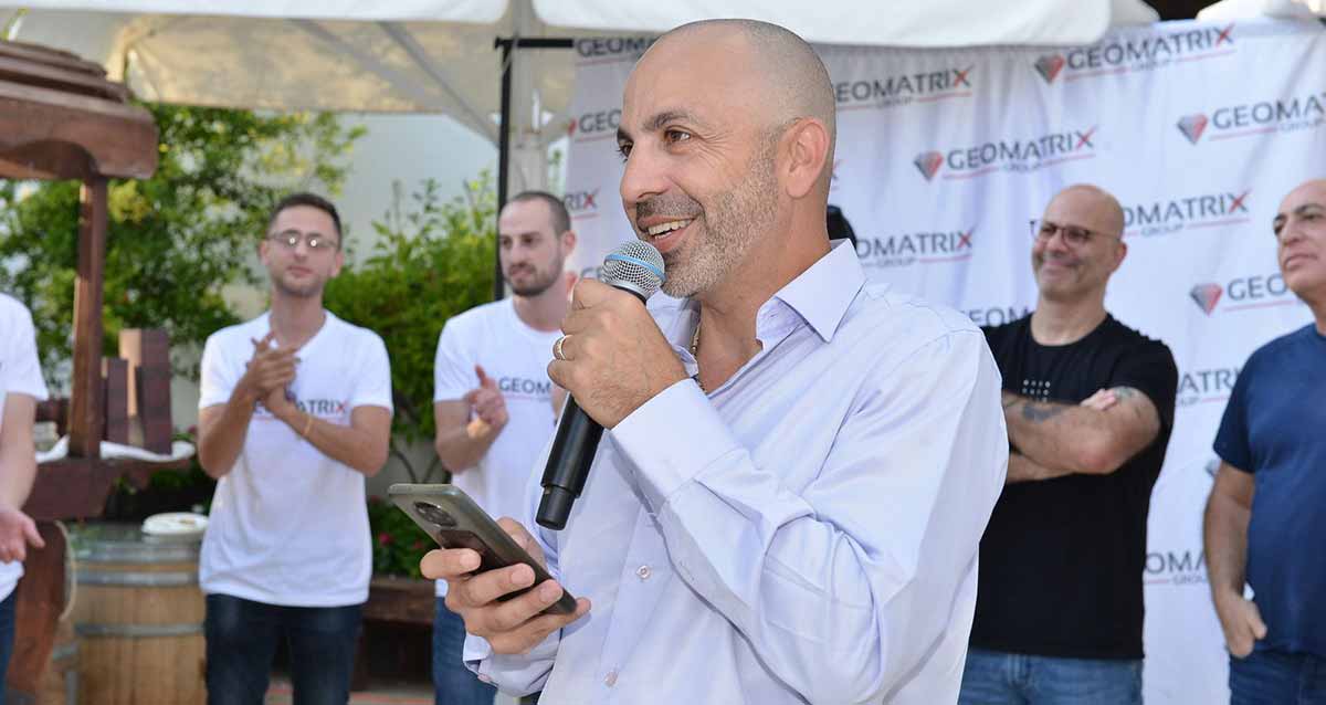 צוות גיאומטריקס רעננה. אדם עם מיקרופון וטלפון ביד עומד לפני קבוצה של אנשים. מאחוריהם על הקיר יריעת בד עם סמל החברה ושמה באנגלית: GEOMATRIX.