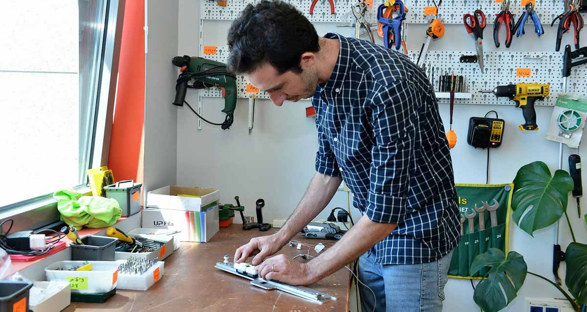 צוות גיאומטריקס רעננה. עובד החברה עומד ליד שולחן עבודה ומטפל במכשיר ארוך. על השולחן קופסאות ועל הקיר מאחוריו תלויים כלי עבודה.