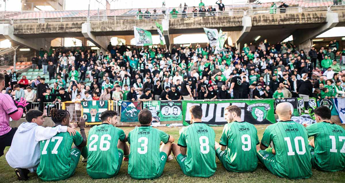 הפועל 1928 כפר סבא. השחקנים בתלבושת ירוקה יושבים על הדשא מול יציע הקהל.