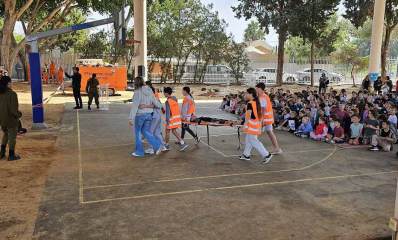 תרגיל חירום בבית הספר היסודי גש"ר בכפר שמריהו. במגרש הספורט ארבעה תלמידים נושאים אלונקה. ברקע גרר כתום של יחידת חילוץ והצלה, ומספר מבוגרים בצד שמאל. מימין יושבים על המשטח התלמידים הצופים בתרגיל.