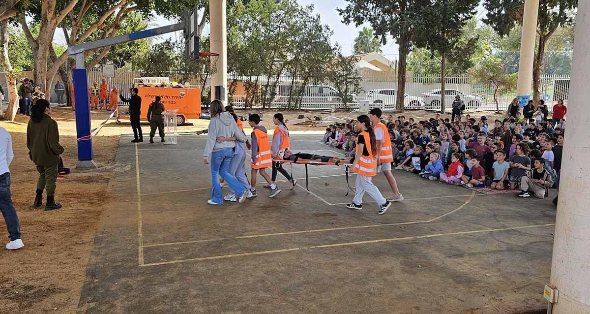 תרגיל חירום בבית הספר היסודי גש"ר בכפר שמריהו. במגרש הספורט ארבעה תלמידים נושאים אלונקה. ברקע גרר כתום של יחידת חילוץ והצלה, ומספר מבוגרים בצד שמאל. מימין יושבים על המשטח התלמידים הצופים בתרגיל.
