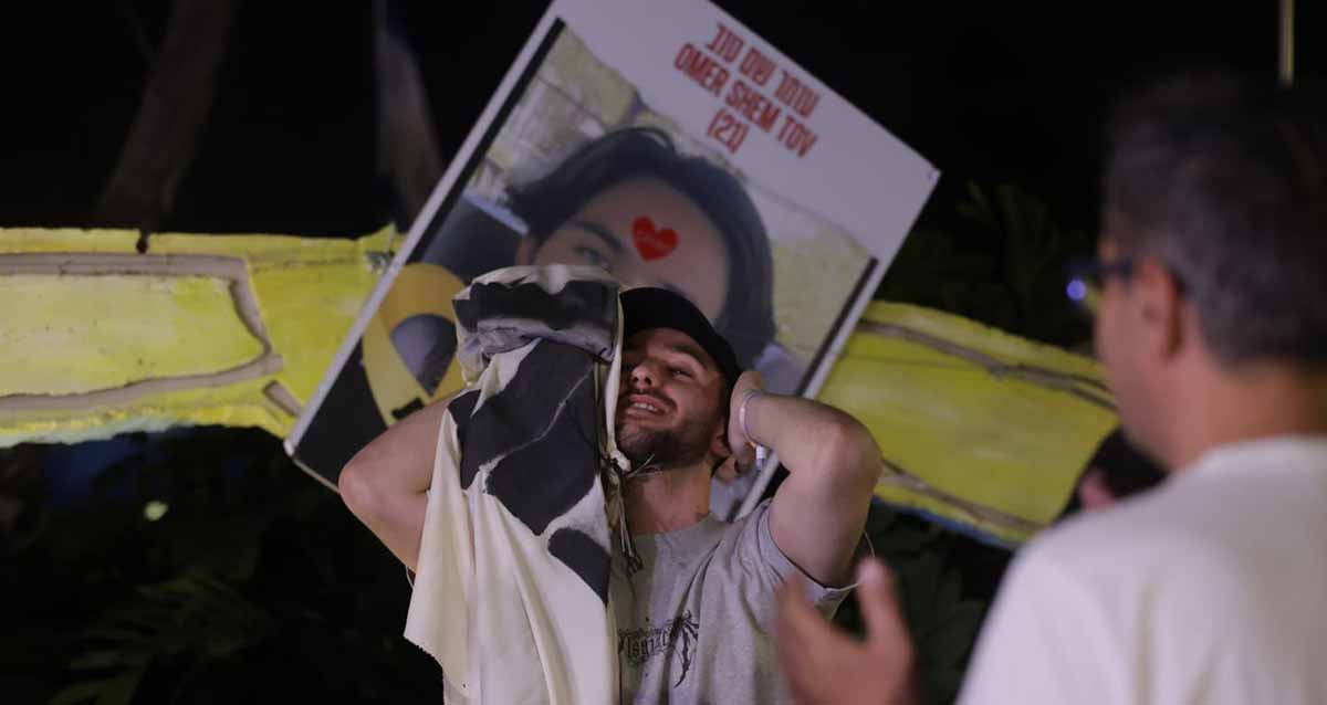 עומר שם טוב עם ידיים על הראש. מאחוריו הפלקט שפורסם לפני שחזר מהשבי.