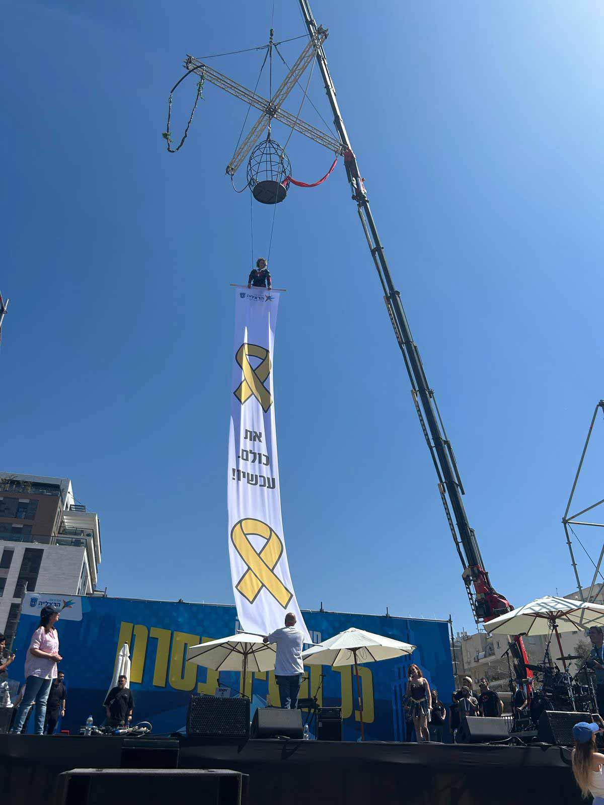 אירוע פורים המרכזי בהרצליה. מנוף מרים שלט בד ארוך מעל במת הבידור, ועליו שני הסמלים הצהובים של החטופים וביניהם הכיתוב: את כולם עכשיו! על הבמה ברקע כיתוב: כל העיר בסרט.