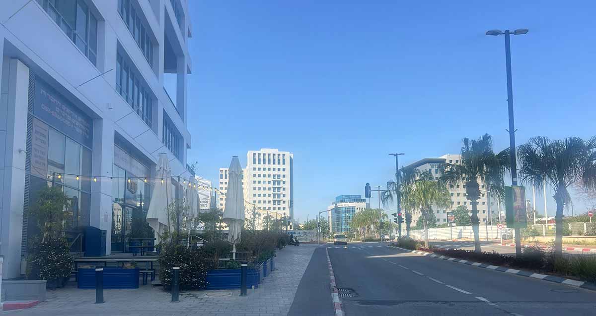 אזור התעשייה רעננה. בצד שמאל מבנה בן ארבע קומות. בית קפה בקומת הקרקע ואזור ישיבה עם שמשמיות במדרכה הרחבה. ברקע מבנים גבוהים.