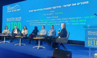 רוכברגר בכנס רשות מקרקעי ישראל. על הבמה יושבות 4 נשים ו-3 גברים ולפניהם שולחנות עגולים. מאחוריהם על הקיר מוקרן הכיתוב: מחברים את ישראל - פרוייקטי התחבורה הגדולים של ישראל