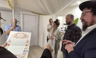 חתונה במועצה הדתית רמת השרון. חתן וכלה מתחת לחופה. רב מקריא את הכתובה. לידו עוזר. אנשים אוחזים את עמודי החופה.