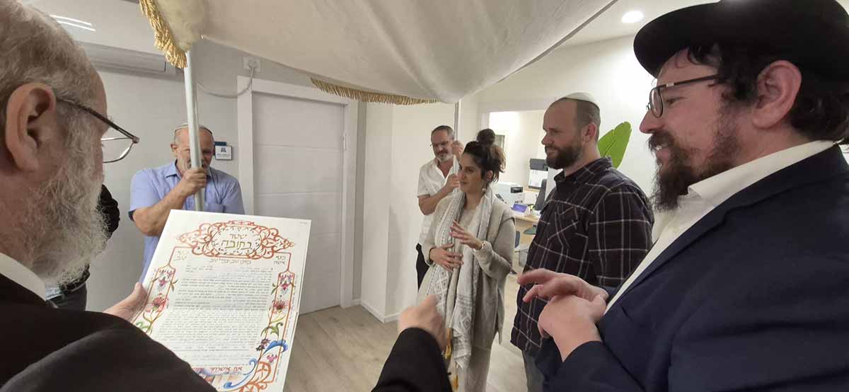 חתונה במועצה הדתית רמת השרון. חתן וכלה מתחת לחופה. רב מקריא את הכתובה. לידו עוזר. אנשים אוחזים את עמודי החופה.