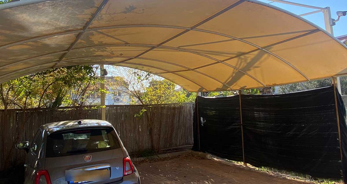 סככה קשתית לבנה וחניית עפר מוקפת בגדר במבוק וגדר בדי יוטה. בחניה מכונית אפורה. בצד ימין על הקונסטרוקציה של הסככה מוצבת מצלמת אבטחה
