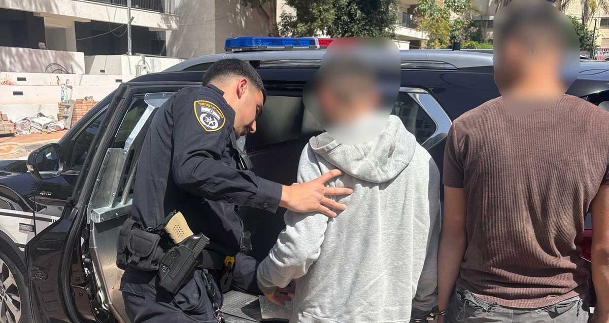 שוטר עוצר שני שוהים בלתי חוקיים ליד אתר בנייה. לפניו ניידת משטרה