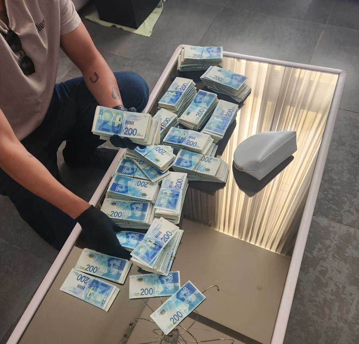 מזומנים שנתפסו, חבילות של שטרות 200 שקלים על שולחן, ושוטר עם כפפות שחורות מטפל בהן.