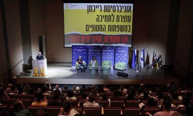על קיר הבמה מוקרן הכיתוב: אוניברסיטת רייכמן עוצרת לתמיכה במשפחות החטופים, על הבמה ארבעה כסאות ושניים יושבים, משמאל דוכן נואמים עם אדם לידו. בצד ימין יושב נגן גיטרה ולידו דגלי המדינה. באולם יושב הקהל.