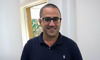 עו"ד טל שלומי מרכיב משקפי ראייה, בטישרט בצבע כחול כהה. ברקע דלת המשרד ותמונה בקיר המשרד.