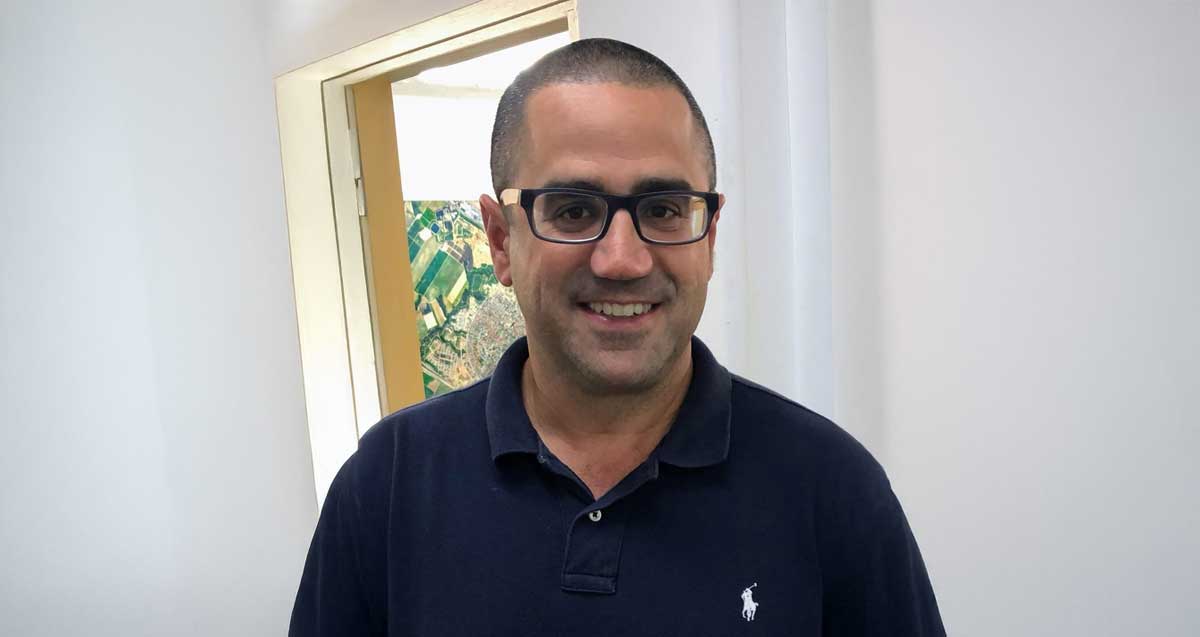 עו"ד טל שלומי מרכיב משקפי ראייה, בטישרט בצבע כחול כהה. ברקע דלת המשרד ותמונה בקיר המשרד.