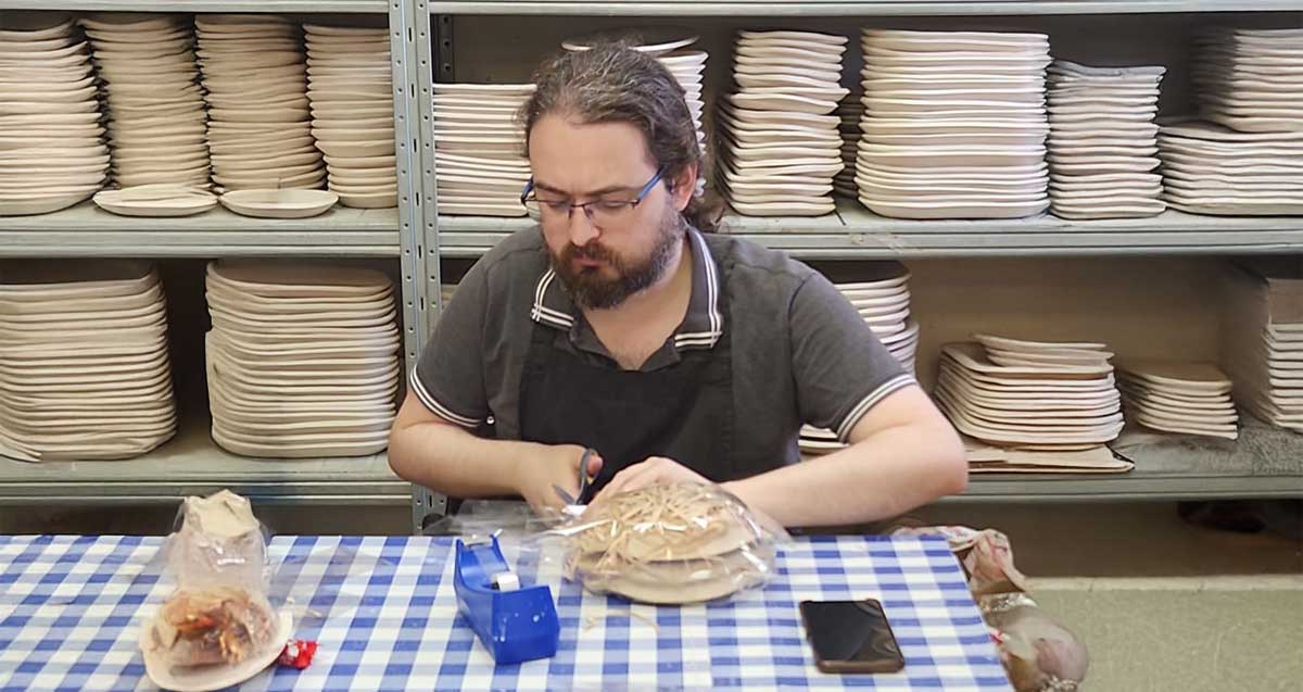 עמית רחמימוב יושב ליד שולחן עם מפה בצבעים כחול ולבן, וסדר אריזה העטופה בפלסטיק שקוף. על השולחן מתקן עם צלוטייפ. מאחוריו לאורך הקיר מדפי מתכת עם צלחות בגדלים שונים.