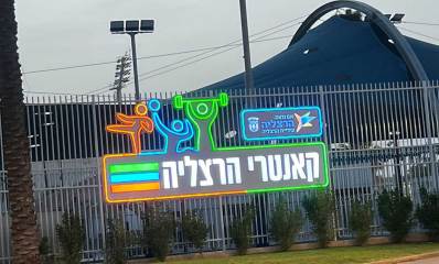 קאנטרי קלאב עירוני הרצליה. צילום מבחוץ. אוהל כחול מעל המועדון. על הגדר שלט עם הכיתוב: אם נרצה. הרצליה, עיריית הרצליה, ושלט גדול עם הכיתוב: קאנטרי הרצליה. מעל השלט הגדול דמויות של ספורטאים משלושה ענפי ספורט.