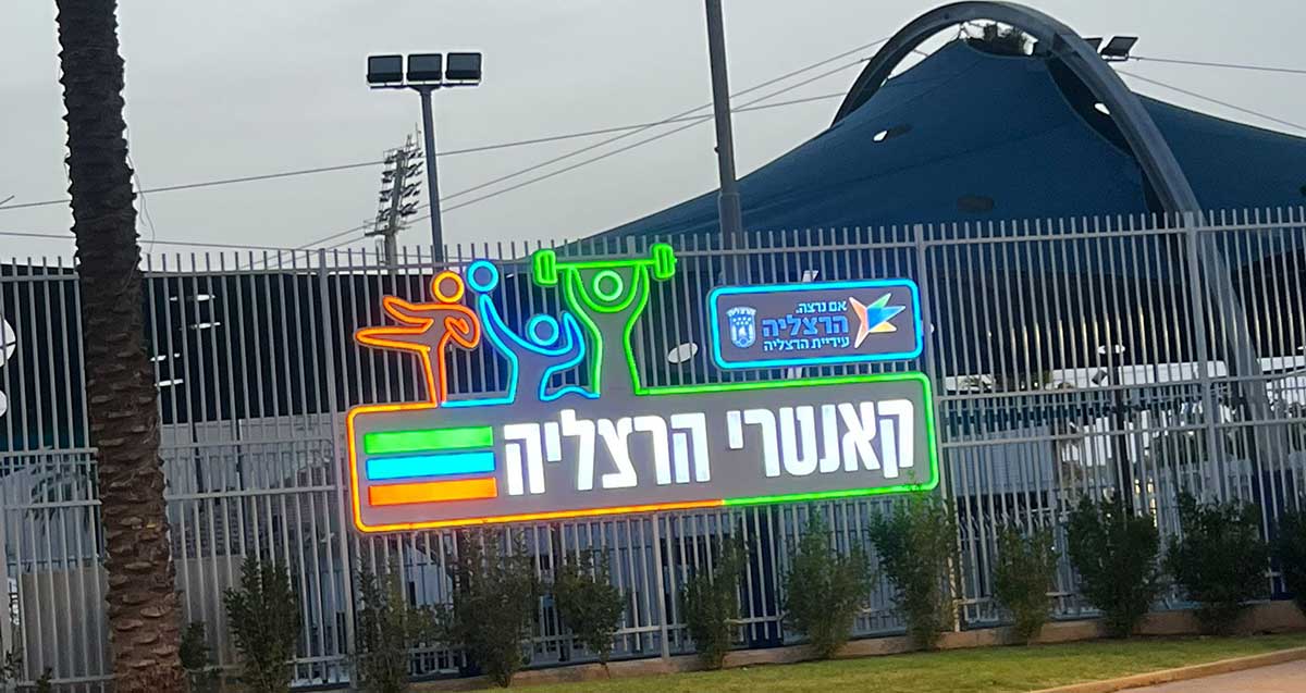 קאנטרי קלאב עירוני הרצליה. צילום מבחוץ. אוהל כחול מעל המועדון. על הגדר שלט עם הכיתוב: אם נרצה. הרצליה, עיריית הרצליה, ושלט גדול עם הכיתוב: קאנטרי הרצליה. מעל השלט הגדול דמויות של ספורטאים משלושה ענפי ספורט.