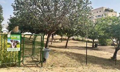 גינת הכלבים הזמנית בחורשת החקלאים בשכונת נווה זמר. גינה של חול עם עצים. בכניסה שלט עם תמונה של ראש כלב וכיתוב: הוא עשה את שלו, ואתם? רעננה נקייה