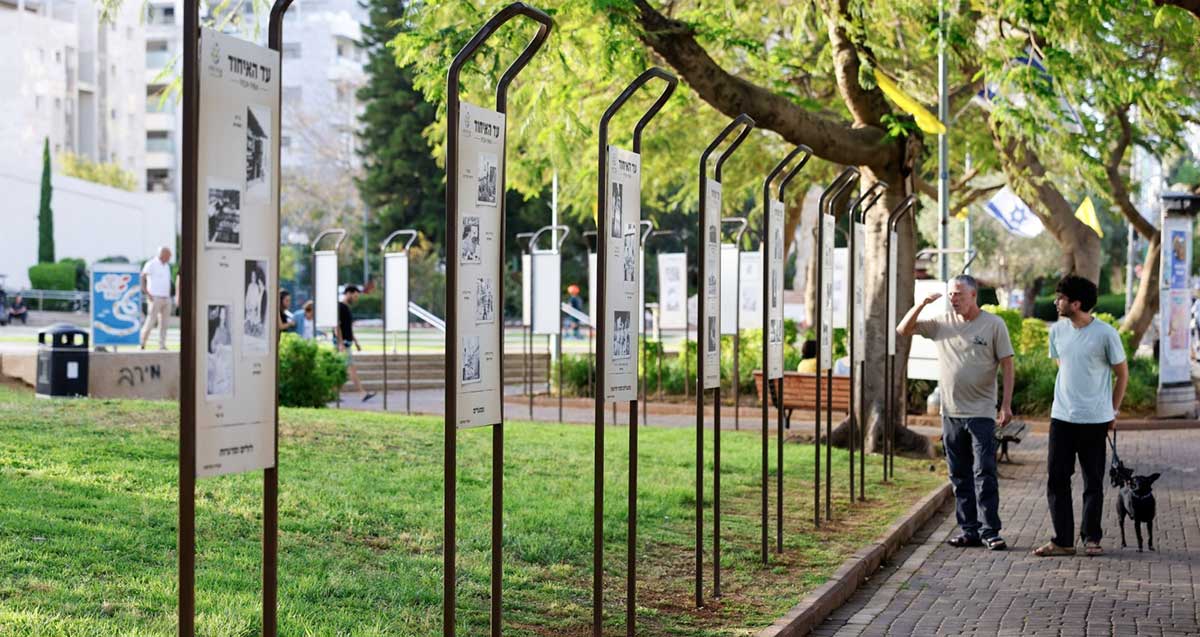 תערוכת "עד האיחוד" בפארק. שורה של שלטים ארוכים וגבוהים, עם כותרת בכיתוב: עד האיחוד, ובכל שלט שלוש או ארבע תמונות. שני אנשים בשביל של הפארק עומדים וצופים באחד השלטים.