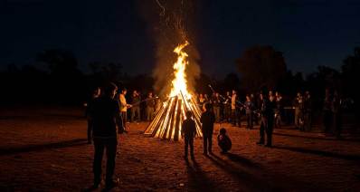 מדורת ל"ג בעומר בצורת קונוס עם אש חזקה וסביבה עומדים ילדים ומבוגרים. המבוגרים הקרובים למדורה אוחזים מקל שקצהו בוער.
