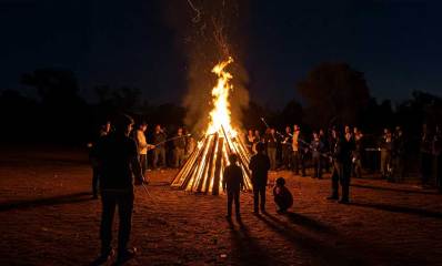 מדורת ל"ג בעומר בצורת קונוס עם אש חזקה וסביבה עומדים ילדים ומבוגרים. המבוגרים הקרובים למדורה אוחזים מקל שקצהו בוער.