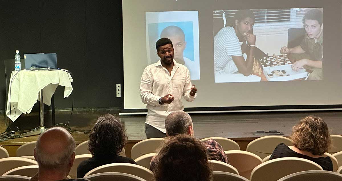 משה וורקו בהרצאה בבית יד לבנים ברעננה, עומד לפני הקהל היושב על כיסאות. על מסך מאחוריו מוקרנות שתי תמונות, אחת שלו והשנייה הוא משחק שחמט עם מישהו. בצד שמאל על הבמה מחשב נייד ובקבוק מים עם שולחן עגול.