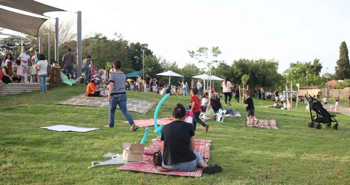 פארק רבין בשכונת נווה עמל. מרחב גדול עם דשא ושמשיות, ומשפחות יושבות על שטיחים ויריעות בד.