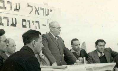 אברהם קרן נואם בהנחת אבן פינה לשיכון של חיילים משוחררים ופועלים חקלאים (שיכון למפרט) ב-1947. מעליו על הקיר פלקט עם כיתוב חלקי: ישראל ערבים...