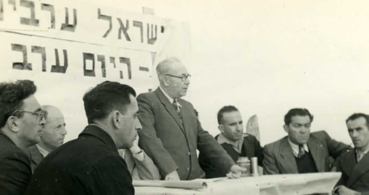 אברהם קרן נואם בהנחת אבן פינה לשיכון של חיילים משוחררים ופועלים חקלאים (שיכון למפרט) ב-1947. מעליו על הקיר פלקט עם כיתוב חלקי: ישראל ערבים...