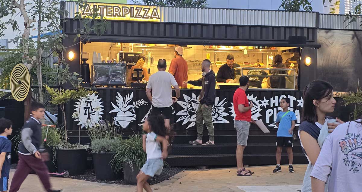 הפיצריה ליד המרכז הקהילתי מטאור ברחוב נתן אלתרמן הרצליה. לקוחות ליד המדרגות של הפיצריה. מעל העסק שלט באנגלית: ALTERPIZZA