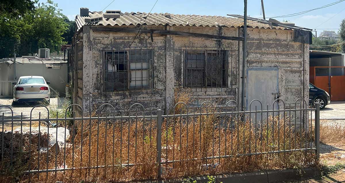 צריף האסבסט ברחוב דוד אלקלעי בשכונת נווה ישראל. דלת ושני חלונות מסורגים בחזית. גג אסבסט משופע.