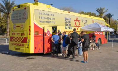 התרמות דם ביום שישי. ניידת תרומת דם של מגן דוד אדום חונה ברחבה, ואנשים עומדים בתור ליד הדלת כדי להיכנס ולתרום. ליד הניידת הוקם מקום מוצל עם כיסאות.