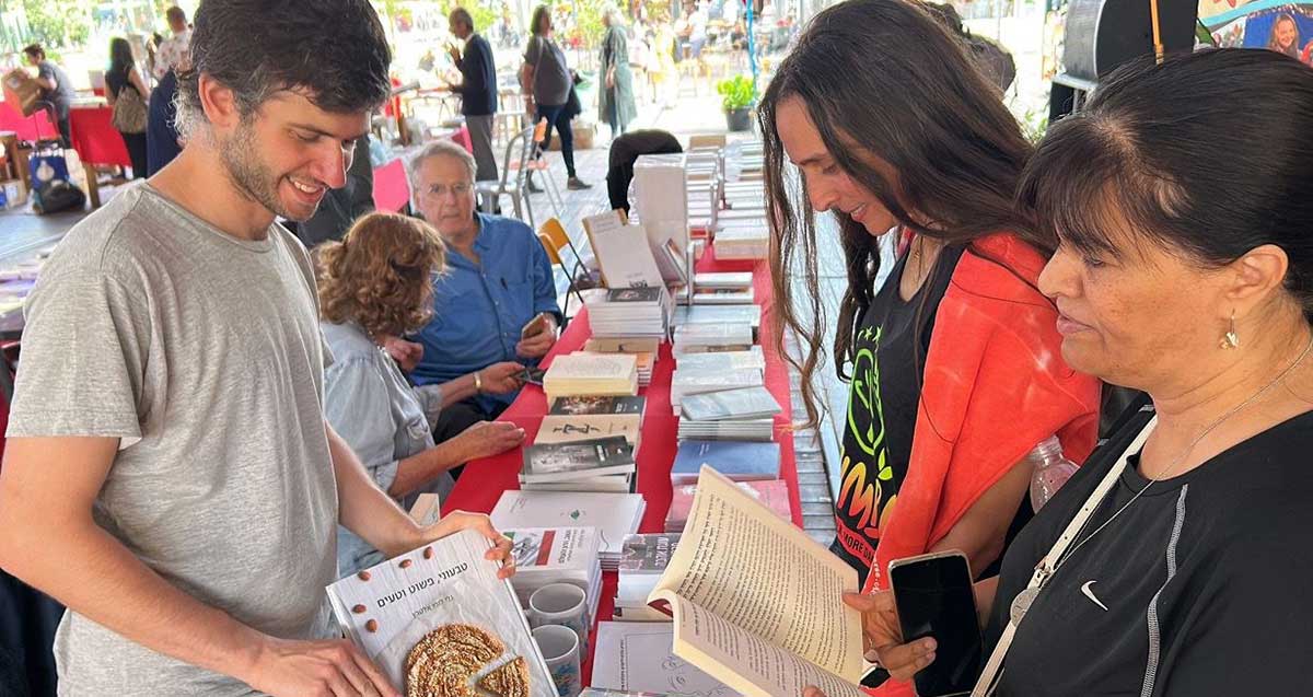 שבוע הספר. שולחן ארוך עם ספרים על מפה אדומה, וקהל ליד השולחן מעיין בספרים. בצד שמאל של השולחן יושבים שניים. ברקע שולחן וקהל בשולחן בכיוון ניצב.