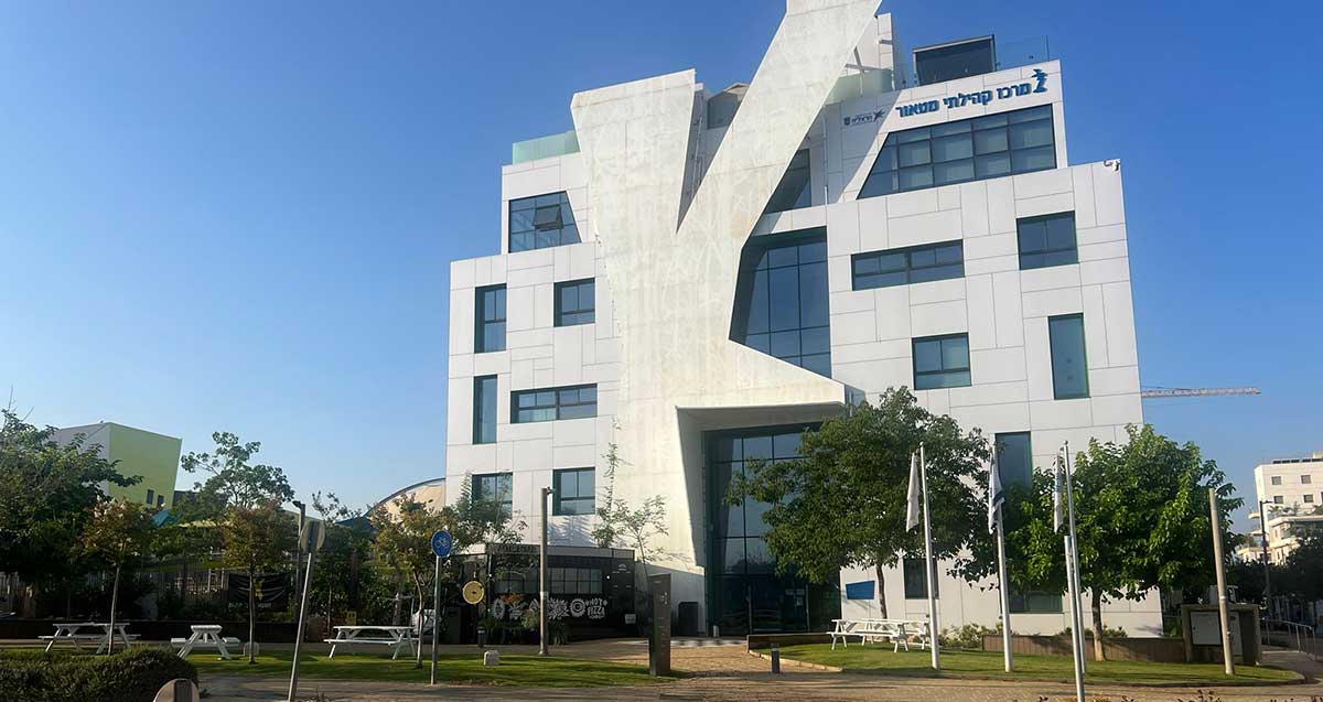 מרכז קהילתי מטאור בהרצליה, הפודטראק בכניסה הראשית. בניין בחיפוי אריחים לבנים. בראש הבניין כתובת: מרכז קהילתי מטאור. לפני הבניין גינה עם שולחנות וספסלים לבנים.