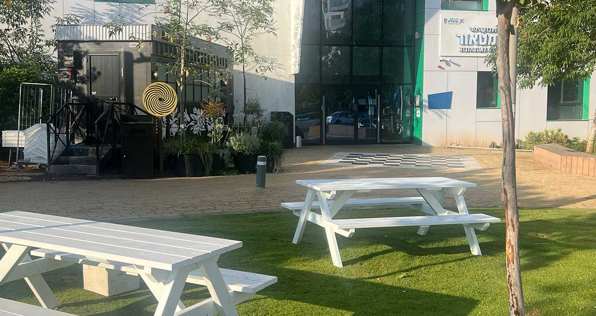 מרכז קהילתי מטאור בהרצליה, הפודטראק בכניסה הראשית בצד שמאל. מעל הכניסה לבניין הכתובת: מרכז קהילתי מטאור ע"ש לאה גולדברג. לפני הבניין גינה עם שולחנות וספסלים לבנים.