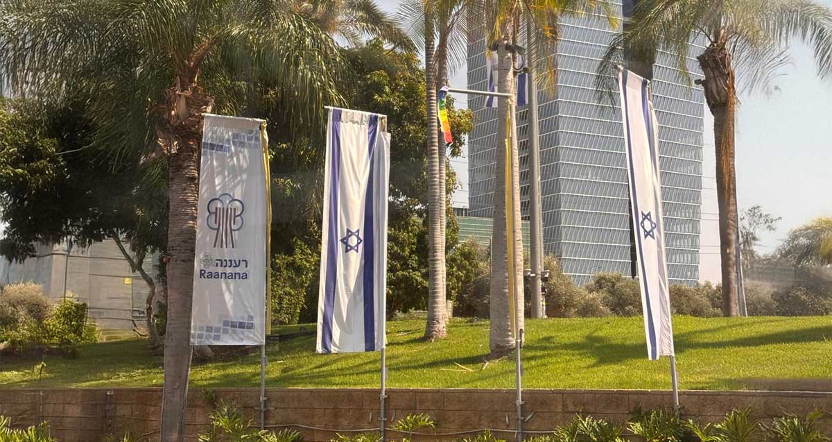 דגל הגאווה שהושחת ברחוב אחוזה ברעננה. מימינו ושמאלו דגלי המדינה ודגל העיר רעננה. הדגלים בחזית של גינה גדולה עם דשא ודקלים. ברקע מגדלים