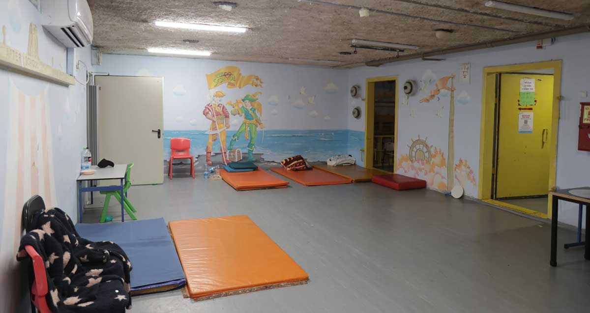 מקלט ציבורי בבית ספר. על הרצפה מונחים מזרונים, כסאות ושולחן. ציורים על קירות המקלט.