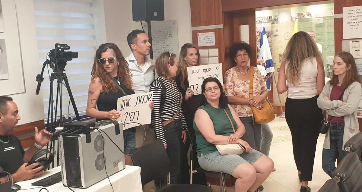 מחאת תושבי שכונת סביוני הכפר בישיבת מועצת עיריית כפר סבא. קבוצה של נשים וגבר עומדים בפתח חדר הישיבות. שתי נשים אוחזות שלטים. שלט עם כיתוב: שכחת אותנו רפי?! שלט שני: ילדי שכונת 'סביוני הכפר' שווים פחות ??!