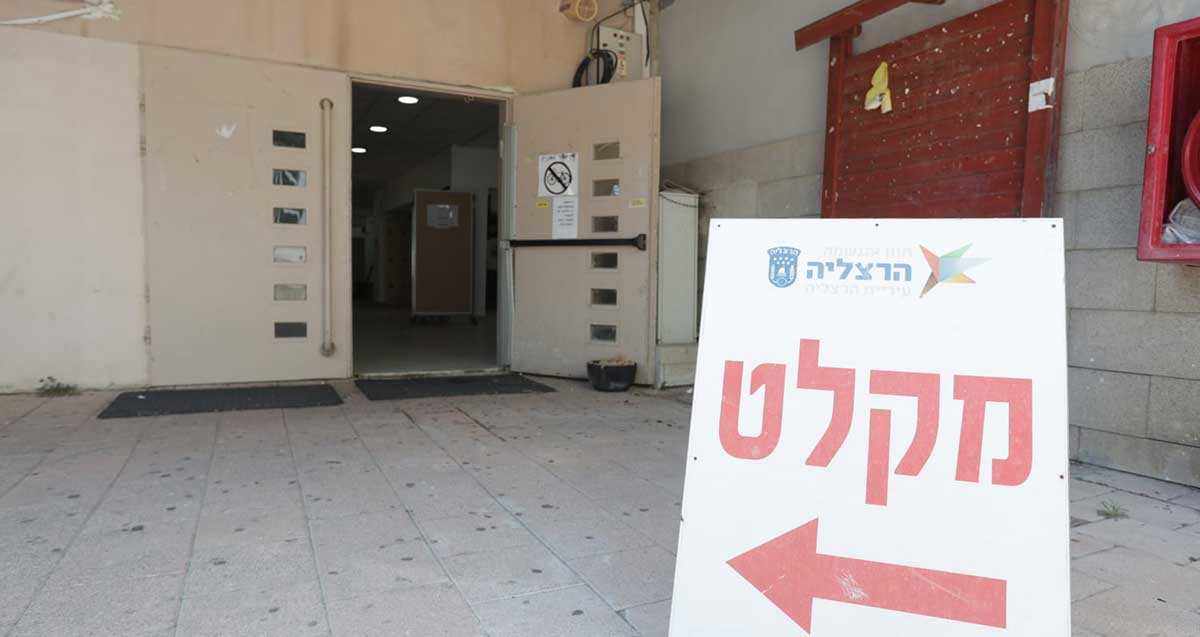 מקלטים בבתי ספר. בבית הספר, שלט הכוונה גדול של העירייה עם חץ אדום וכיתוב: מקלט. ברקע דלת מתכת כפולה פתוחה למחצה.
