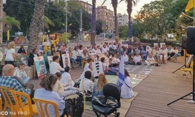 פעילות למען החטופים בכפר סבא. קהל רב יושב ברחבה על כיסאות ועל הרצפה מול מסך ורמקולים.