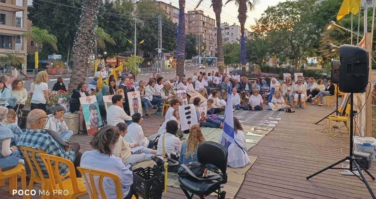 פעילות למען החטופים בכפר סבא. קהל רב יושב ברחבה על כיסאות ועל הרצפה מול מסך ורמקולים.